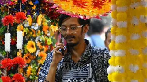NurPhoto via Getty Images A person surrounded by plastic flower decorations talks on his mobile phone while buying home decor ahead of the Diwali festival in Kolkata city on October 19, 2025. 