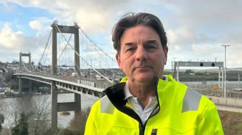 A close up picture of Philip Robinson from Tamar Crossings stood on a balcony overlooking the Tamar Bridge. He is looking straight at the camera and is wearing a high viz coloured coat. 