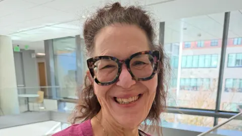 Shekinah Samayah-Thomas Shekinah Samayah-Thomas, wearing thick tortoiseshell glasses, smiling broadly in what appears to be an office building, with floor-to-ceiling windows behind her