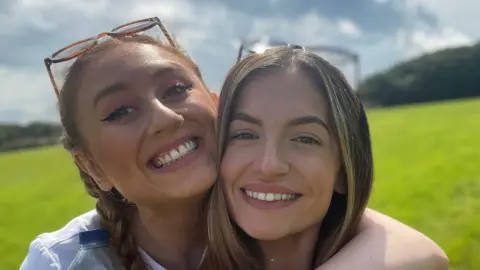 Two women, Daisy and Georgia, smile while embracing - they have sunglasses on their heads and are standing in a field