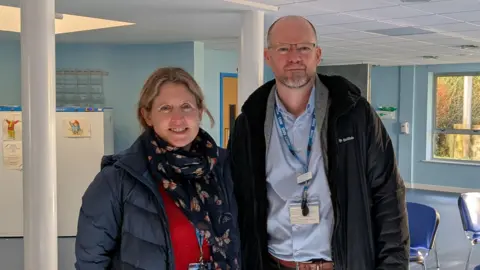 Dr Jennie Button, GP stands on the left wearing a blue scarf and jacket with a red jump, black skirt and tights and a lanyard, while Chris Cruise, practice manager, on the left, wears glasses a pale blue shirt, dark coat and brown trousers, with a lanyard around his neck. Behind them is a waiting room with blue chairs and foliage seen outside through the windows. 