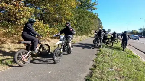 YouTube A group of riders mostly dressed in black, some with helmets and face coverings. They are on a grass verge and path next to a busy road, but appear to be stopped and are facing in different directions.
