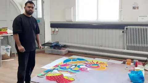 A man all in black standing next to a large rangoli artwork being made inside a hall