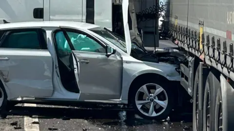 A silver car with its bonnet partially wedged beneath a lorry trailer. Its front is crumpled and a door is open. The air bags have gone off and there is debris on the road surface.