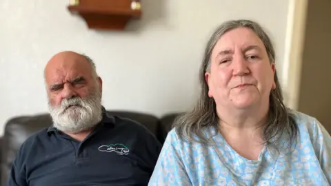 Kevin (left) with receding grey and beard sits on a black leather sofa wearing a navy blue polo top behind Alison who has long grey hair wearing a grey and pale blue patterned top.