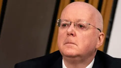 Peter Murrell, who is bald with glasses, in a close-up shot. He is wearing a dark suit, white shirt. 