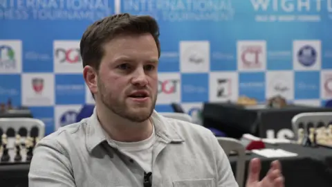 David Lightfoot, a man with neat hair and a short beard, is pictured in conversation at the Isle of Wight Chess Tournament. Tables and boards can be seen behind him. He's wearing a light-coloured collared shirt over a white T-shirt.