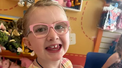 supplied A sweet girl in pink glasses and blonde hair tied back smiles to the camera