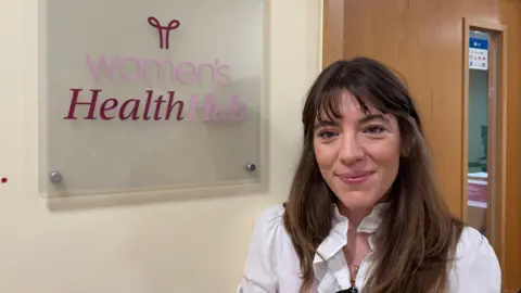 Dr Jessica Langtree-Marsh is smiling at the camera in a heads-and-shoulders photo. She has long brown hair and is wearing a white top. She is standing next to a sign in a corridor that says "Women's Health Hub". There is a door behind her.