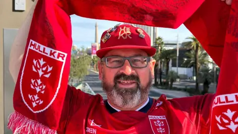 Leonard Gregory Leonard Gregory in Las Vegas. He has a grey and black beard and wears glasses a red Hull Kingston Rovers cap and red team shirt and is holding a club scarf above his head.
