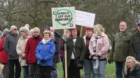 A group of people are taking part in a protest against development on green belt land. A woman is holding a sign which says 'hands off our greenbelt' and has handprints in grey and green paint.
