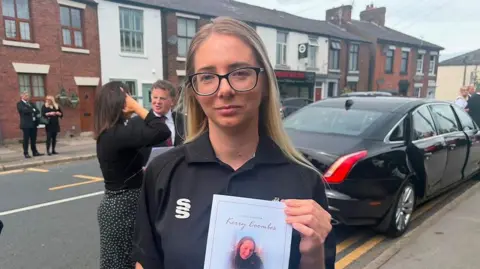 Sinead Garvin with long blond hair wearing black glasses and a black football shirt. She is holding Kerry Coombes funeral order of service. She stands in front of a black vehicle parked outside a church with fellow mourners in the background.