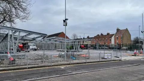 A large construction site where metal frames are being erected. The site is surrounded by a metal security fence. A row of redbrick houses is visible in the background. 