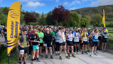 BBC Hundreds of people dressed in running gear lined up at the start line of The Big Cheese race. There are two yellow flags at the start point with the Cheddar Running Club logo.