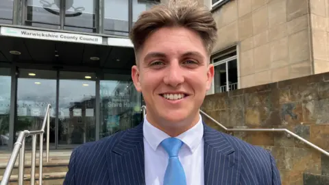 George Finch faces the camera, he has a centre parting in brown hair. He stands outside a building of brown stone in front of stone steps with a metal guardrail.