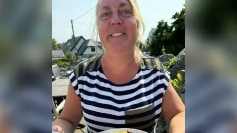 Emma Williams smiles as she sits at a table outdoors, with a plate of food in front of her and a table seen behind her. She has long blonde hair which is tied back and is wearing a black or navy blue and white striped top.