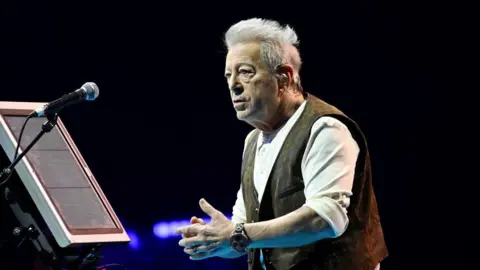 Dave J Hogan/Getty Images A man with wavy white short hair conducting a choir. He is wearing a brown gilet and a white shirt. There is a microphone in front of him and a screen.