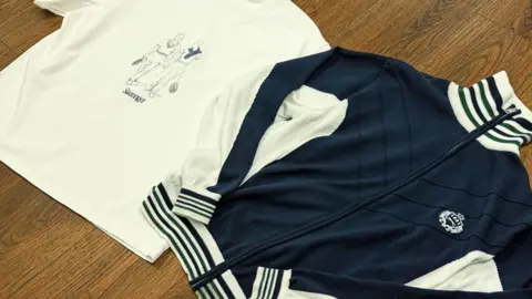 Handout A blue and white jacket is lying on a wooden table alongside a white t-shirt which has a design of two people walking on it. 