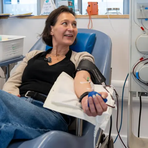 NHS Blood and Transplant Rebecca sits in a medical chair at a plasma donor centre, smiling as she donates plasma, with a needle in her arm and tubing connected to a machine beside her.