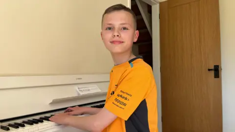 Harvey, 11, sits at a white piano with his hands on the keys and looks at the camera. He is wearing a gold-coloured replica F1 shirt. 