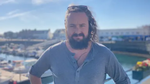 Deputy Lee van Katwyk looking at the camera. There are boats in a harbour behind him. He has long hair and a bushy beard, and has sunglassses on his head.