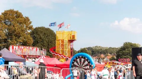 A bright, vibrants image of various rides, slides, and people. There is a helter-skelter towering above the crowds. There is a blue sky and clouds above, and trees in the background.
