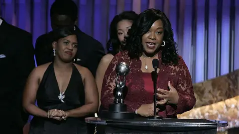 Getty Images Shonda Rhimes, in a dark burgundy lace gown, gives an acceptance speech on stage at an awards show, the Grey's Anatomy cast behind her looking on.