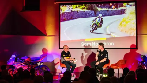 SOUTHERN 100 Naughton being interviewed about his racing at a Southern 100 event. He's sitting opposite event host Chris Kinley on a stage with a screen behind them showing a picture of him racing.