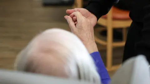 Reuters A view from behind of an older person with grey hair sitting in an armchair and holding out their right hand. It is being held by a person who is standing. 