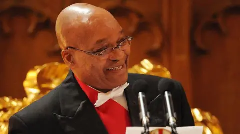 A head and shoulders image of Jacob Zuma in a white bow tie and standing at two microphones. He is looking to his left and is smiling.