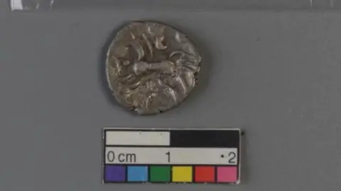 Corinium Museum A close up of an Iron Age coin next to a small sign showing centimetres. The coin is silver in colour and bears the image of a three-tailed horse.