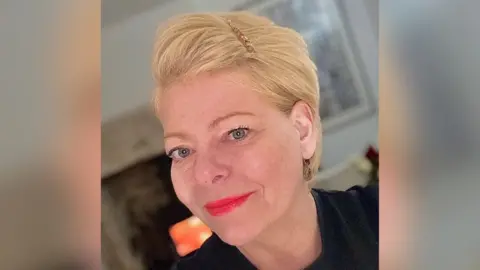 Caroline The image shows a woman with short blonde hair, red lipstick, and a black T‑shirt standing in front of a softly blurred background, with a fireplace visible behind her.