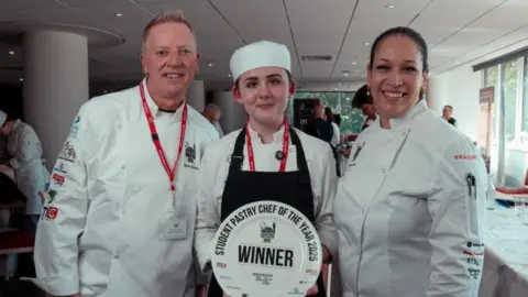 Phoebe Mortimer stood holding a plate-shaped award which says "Winner" and "student pastry chef of the year 2025" on it. She is stood between two people, a man on her right and a woman on her left. They are all wearing chefs whites.