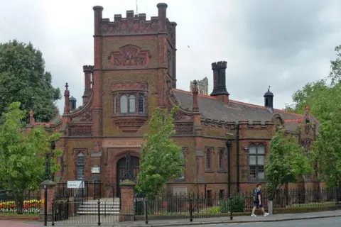 Stephen Richards/Geograph The ornate brick, turretted Carnegie Library on London Road in King's Lynn. The entrance has a tower on top of it, with the building lined with small trees, flowers and iron railings