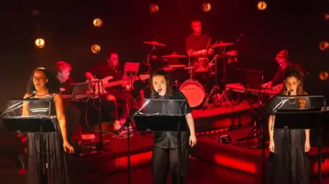 Birmingham Hippodrome/Danny Kaan Three women dressed in black standing on a stage and singing into microphones. Behind them is a band, which is lit up in red. They are Elexi Walker, Maddison Bulleyment and Rosie Coles. 