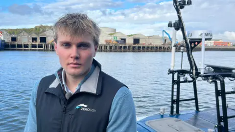 Bertie Ratsey is wearing a light blue top and a navy blue gilet with the company name ZEROUSV and logo and is standing on a pontoon with his unmanned vessel on the water behind him.