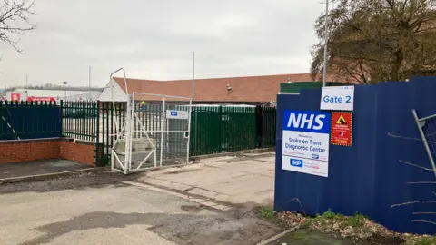 BBC An entrance with a white gate that is open. There is a sign including the letters NHS and Gate 2 on the right. A building is in the background.