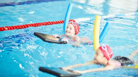 AFP via Getty Images Two schoolgirls in swimwear in a pool, using special swim devices learning to swim.