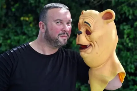 Henry Godfrey-Evans/BBC A man holding up a creepy silicon yellow mask of Winnie-The-Pooh and side-eying the camera. There are bushes behind him.