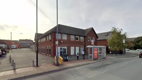 The bank stands on the side of a road behind a bus stop. It is a red-brick building with two levels of windows