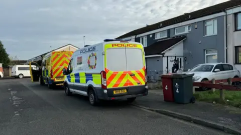 Two Merseyside Police vans parked on a pavement outside a row of terraced houses. A uniformed officer with a black beard stands outside the door of a house with its brickwork painted grey