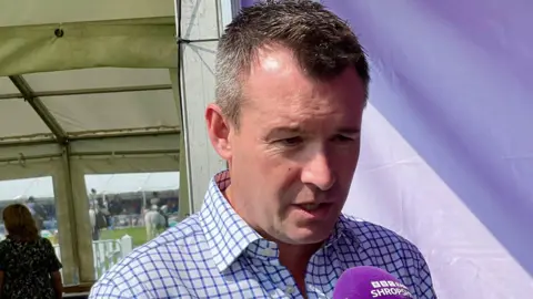 A man wearing a blue checked shirt speaks into a microphone that has a purple cover saying BBC Radio Shropshire