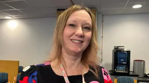 Nicola Haseler/BBC Jody Swain, a teacher who is wearing a predominantly black outside with coloured patterns on it. She has blonde hair and is standing inside a classroom and is looking directly at the camera and smiling.