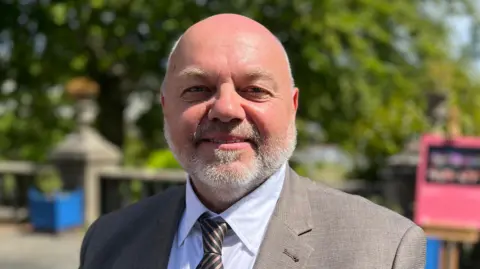 A middle aged bald man with a grey beard in a suit looks at the camera.