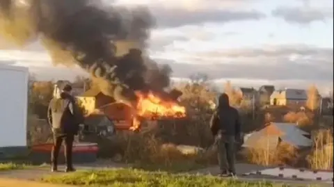 Reuters Grainy image of flames seen rising from debris in a residential area of Russia. Two people can be seen with their backs to the camera, looking at the scene of debris. Thick black smoke is rising from the flames.