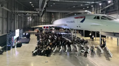 BBC The large hangar at Aerospace Bristol, housing the Concorde aeroplane, with rows of seats and students looking at a temporary stage