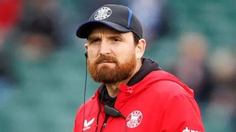 Martin Gleeson standing on the pitch wearing a Bath cap and red team jacket during a warm-up ahead of the Champions Cup match against Saracens at the Recreation Ground