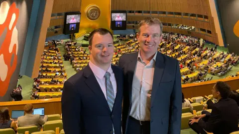 Bekki Cox Two men in suits smile for the camera while standing in a large United Nations hall, where many people are seated