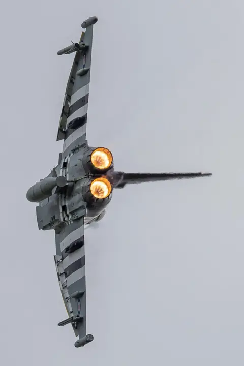 RAF Cosford A streamlined plane flies through the sky on its side. Its afterburners are on and glowing bright orange.