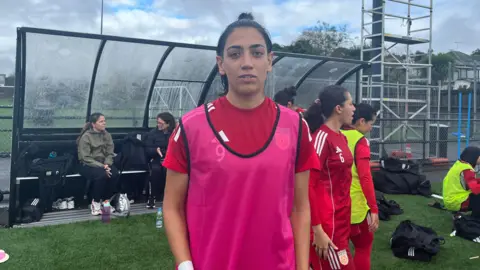 Manozh Noori standing on the edge of a pitch looking at the camera. She has shortish dark hair that is tied back and wears a bright pink vest over her red football kit. In the background there are some kit bags, a few other players and a couple of women sitting in a shelter. The sky is cloudy.
